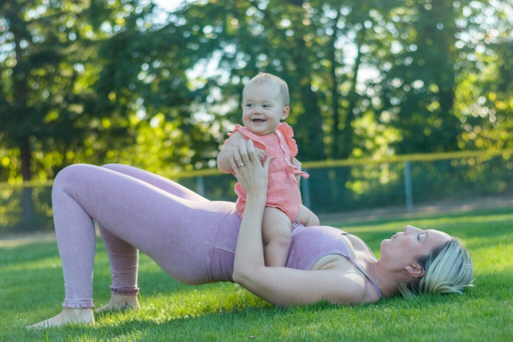 Eine Frau liegt in der Schulterbrücke auf dem Rücken, ein Baby sitzt auf ihrem Bauch und lacht.